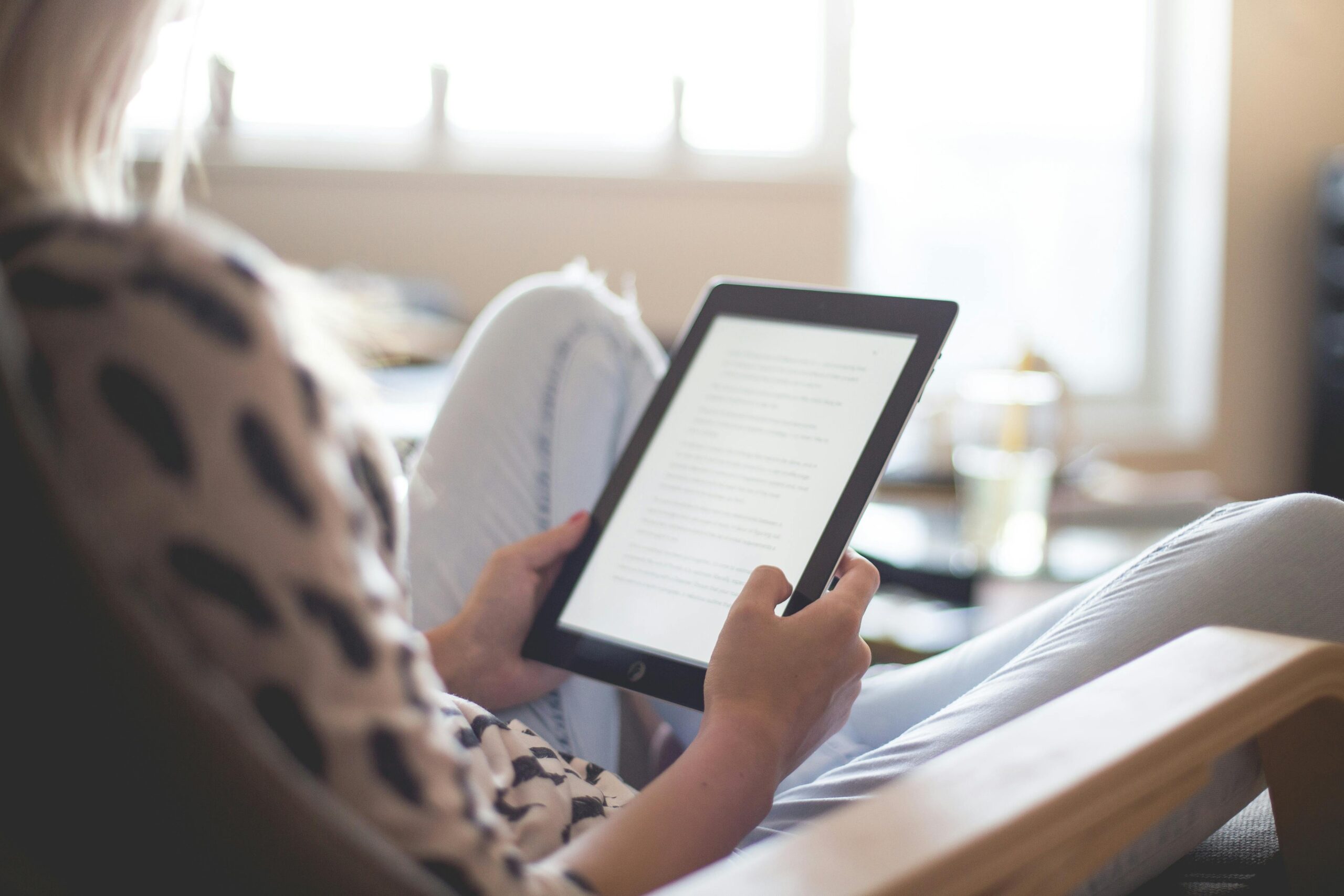 Mujer sentada en una silla leyendo un libro digital en una tablet con luz natural entrando por la ventana